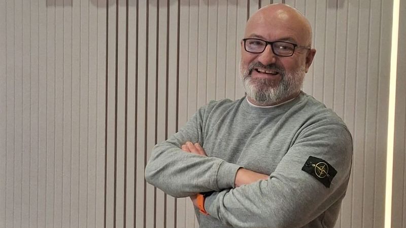 Plano medio de Joan Valls sonriendo con los brazos cruzados, vistiendo sudadera gris y gafas, en un estudio con paneles de madera.