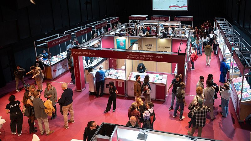Vista cenital de una feria del libro teatral con estands modulares, moqueta fucsia y visitantes ojeando publicaciones en los expositores.