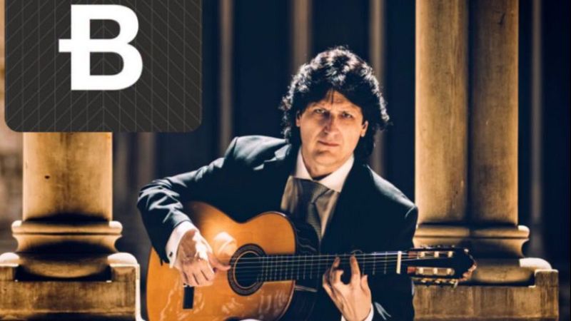 El guitarrista Juan Manuel Ca�izares posa de frente con una guitarra en un claustro de columnas cl�sicas, luciendo traje oscuro y expresi�n seria.