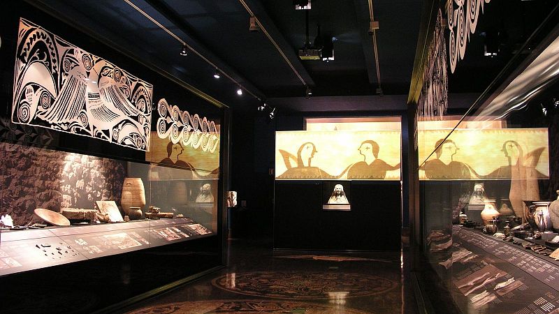 Sala de exposición moderna y oscura con vitrinas iluminadas que muestran vasijas cerámicas y un busto esculpido bajo un panel retroiluminado.