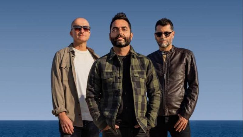 Los tres integrantes del grupo Café Quijano posan juntos al aire libre frente al mar bajo un cielo despejado.