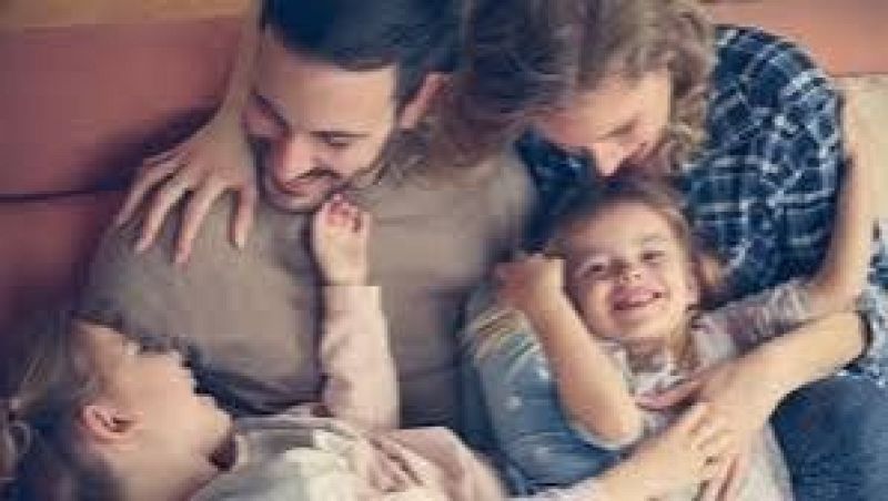 Escena familiar alegre y luminosa donde un padre, una madre y sus dos hijas comparten un momento de juego y complicidad en el hogar.