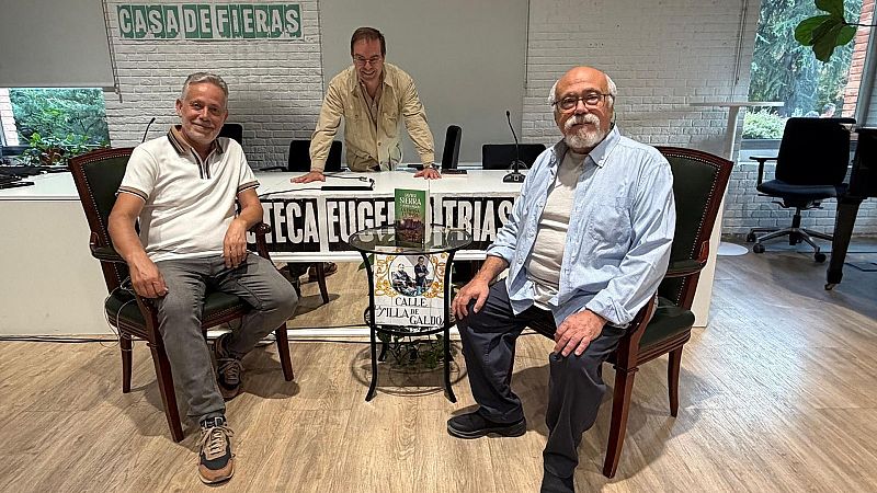 Tres hombres participan en un evento literario en una sala luminosa, sentados a ambos lados de una mesa de ponencias y junto a una mesa auxiliar con un libro y una placa de Gald�s.