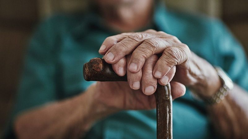 Primer plano de las manos arrugadas de un anciano apoyadas sobre un bastón de madera, con una camisa verde y un reloj.