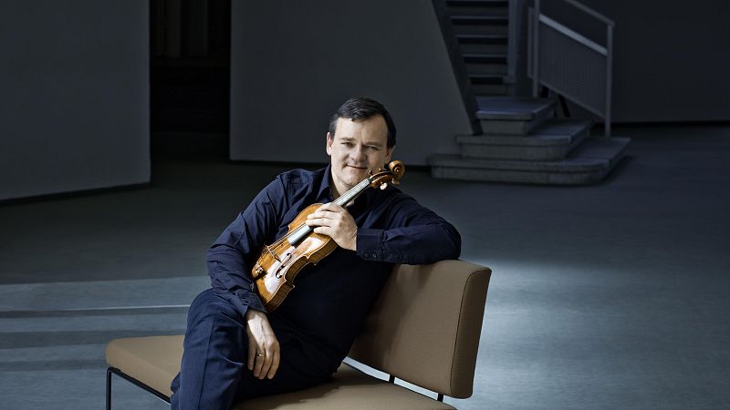 Retrato de un violinista sentado en un banco con un viol�n de madera clara en un espacio moderno y sobrio.