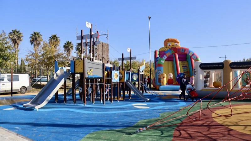 Parque infantil al aire libre con estructura de juegos en forma de barco pirata, toboganes, suelo de caucho de colores y atracciones hinchables al fondo bajo un cielo despejado.