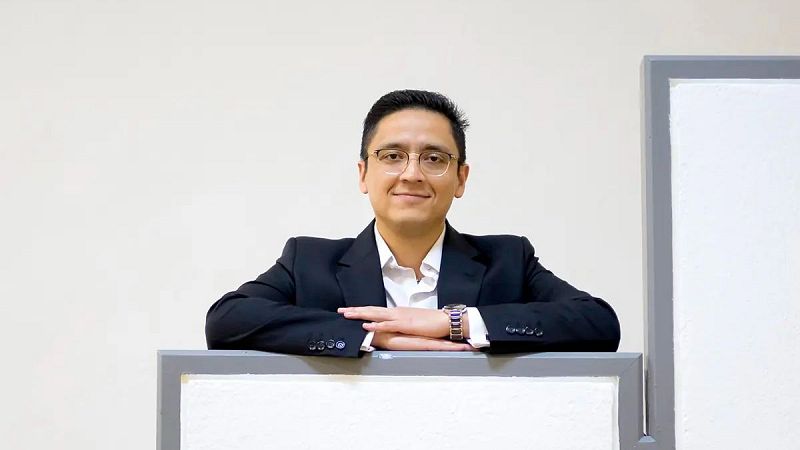 José Eduardo sonríe en un retrato de plano medio con chaqueta oscura, camisa blanca, gafas y reloj metálico ante un fondo minimalista.