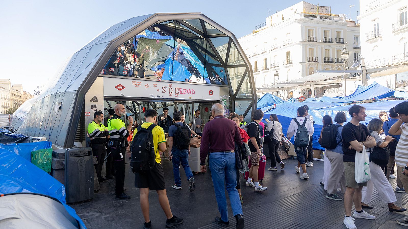 Vista diurna de la Puerta del Sol en Madrid con el pabellón de acceso al metro rodeado por una acampada con lonas azules, agentes de policía y peatones bajo el sol.