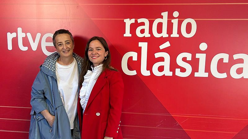 Dos mujeres sonrientes posan ante un panel rojo de RTVE; la de la izquierda lleva el pelo corto y abrigo azul, y la de la derecha una americana roja.