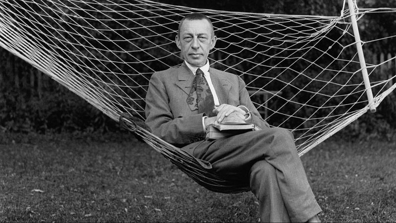 Fotograf�a en blanco y negro del compositor Sergu�i Rajm�ninov sentado en una hamaca al aire libre, vistiendo traje formal y sujetando unos cuadernos.