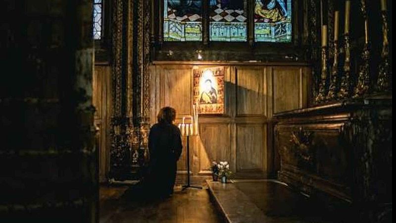 Persona arrodillada frente a un icono religioso iluminado por velas en una capilla oscura con detalles de madera y vidrieras.