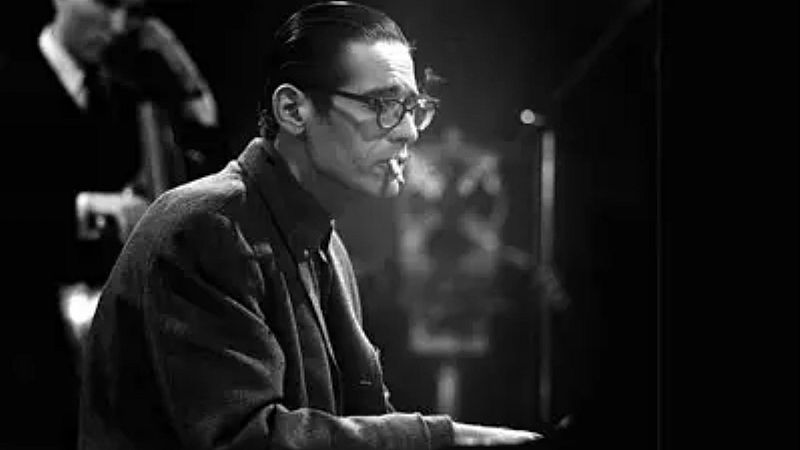 Fotograf�a en blanco y negro de Bill Evans tocando el piano de perfil, con gafas y un cigarrillo encendido, acompa�ado al fondo por un contrabajo.