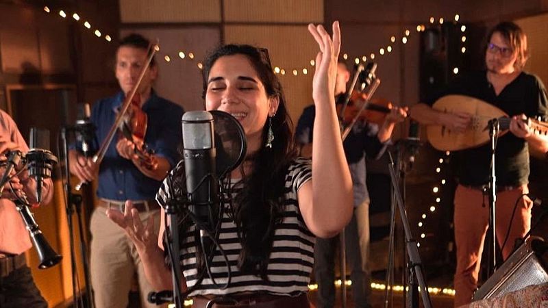 Joven cantante interpreta un tema con pasi�n frente a un micr�fono en un estudio ac�stico decorado con luces c�lidas y acompa�ado por m�sicos con instrumentos de cuerda.