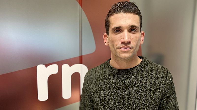 Plano medio corto del actor español Jaime Lorente con jersey verde oliva y expresión serena ante un panel rojo.