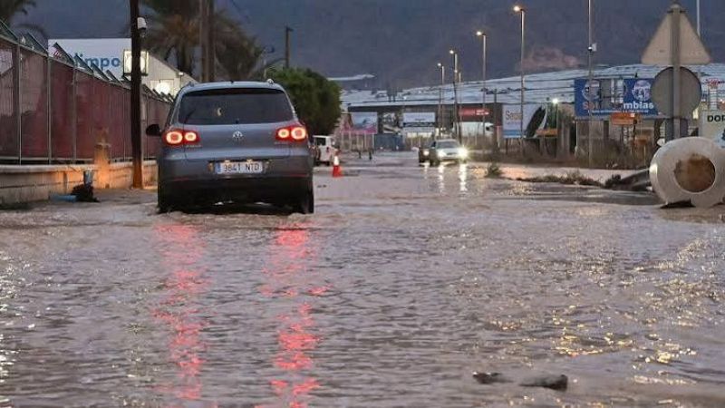 Un todocamino gris circula por una calle anegada por el agua turbia de una inundaci�n, con invernaderos al fondo bajo un cielo gris.