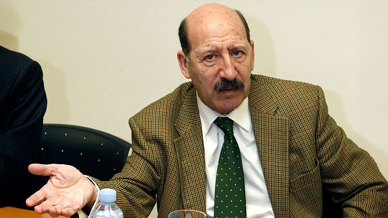 Plano medio del político Javier Sáenz de Cosculluela sentado en una mesa con chaqueta de cuadros y bigote, realizando un gesto explicativo.