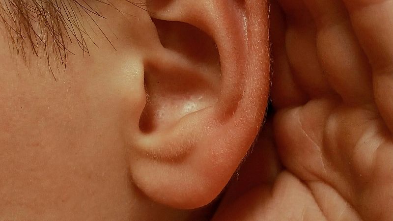 Plano detalle de una oreja humana junto a una mano colocada en gesto de amplificar el sonido, con iluminaci�n suave y c�lida.