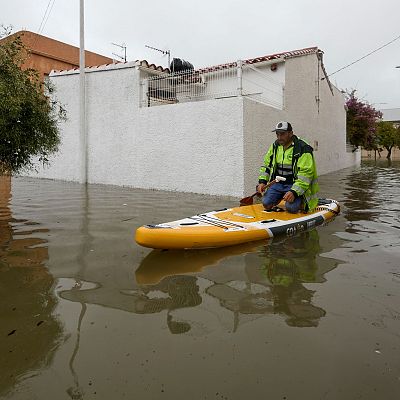 Un hombre con chaqueta reflectante navega de rodillas sobre una tabla de paddle surf en una calle residencial inundada.