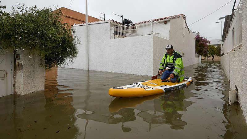 Un hombre con chaqueta reflectante navega de rodillas sobre una tabla de paddle surf en una calle residencial inundada.
