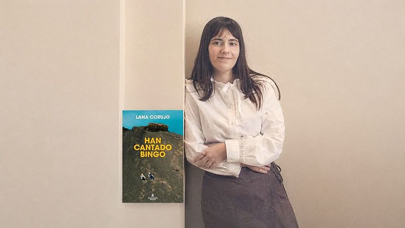La escritora Lana Corujo posa sonriente junto a la portada de su libro 'HAN CANTADO BINGO' en un luminoso espacio interior.