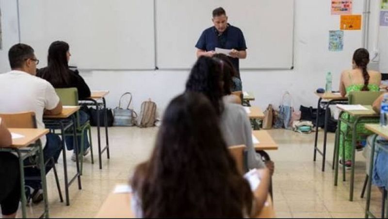 Vista trasera de un aula durante un examen con aspirantes sentados en pupitres y un examinador de pie al frente.