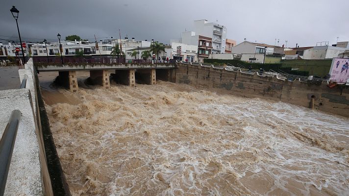 Intensa riada de agua turbia y marrón que discurre con violencia por un canal de hormigón urbano bajo un cielo tormentoso.