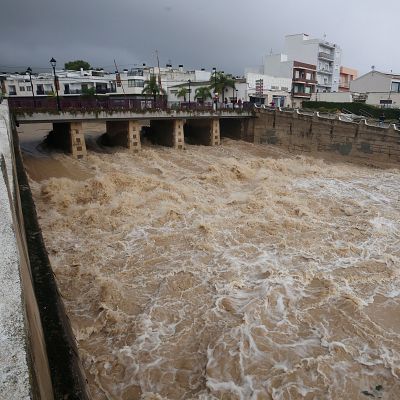 Intensa riada de agua turbia y marrón que discurre con violencia por un canal de hormigón urbano bajo un cielo tormentoso.