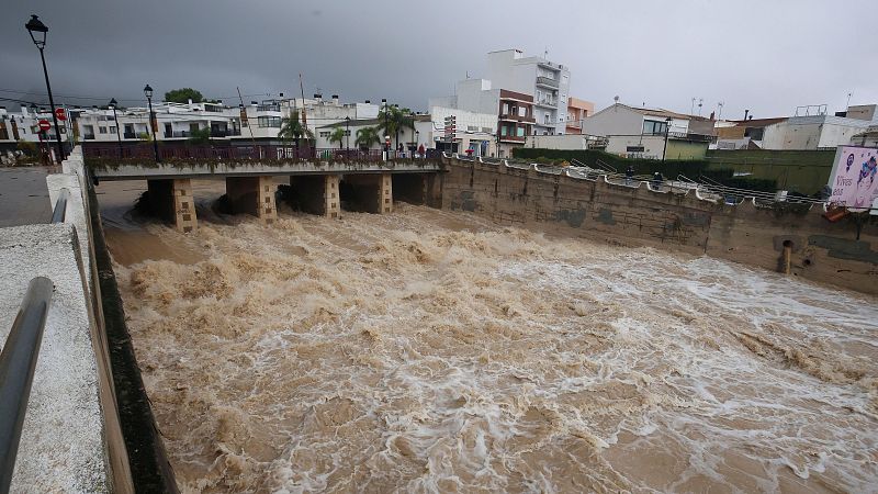 Intensa riada de agua turbia y marrón que discurre con violencia por un canal de hormigón urbano bajo un cielo tormentoso.