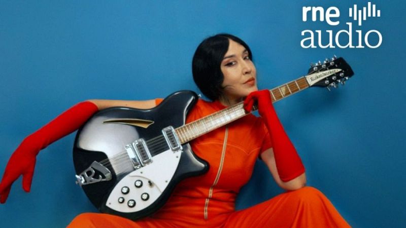La cantante Anni B Sweet con mono naranja y guantes rojos sostiene una guitarra el�ctrica sobre fondo azul, con el logotipo de rne audio.