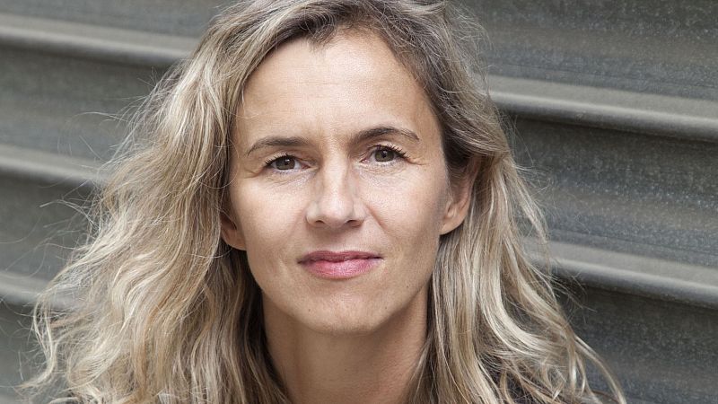 Primer plano de la escritora Delphine de Vigan con expresi�n serena, cabello rubio ondulado y fondo de lamas met�licas grises.