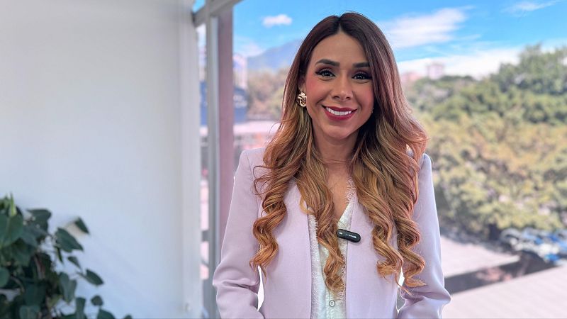 Mujer joven y sonriente con cabello castaño ondulado, chaqueta rosa palo y micrófono en la solapa, en un espacio interior muy luminoso con grandes cristaleras al fondo.