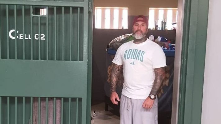 Plano medio de un recluso con brazos tatuados, camiseta blanca y gorra, de pie junto a una puerta de celda metálica numerada como CELL C2.