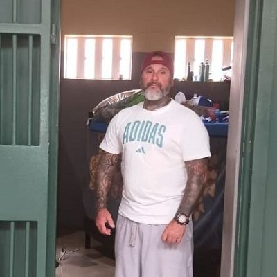 Plano medio de un recluso con brazos tatuados, camiseta blanca y gorra, de pie junto a una puerta de celda metálica numerada como CELL C2.