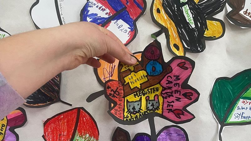 Mano sosteniendo una hoja de papel recortada y coloreada con dibujos y frases infantiles sobre un mural colaborativo.