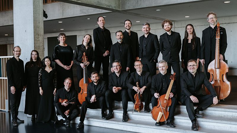 Grupo de diecis�is m�sicos vestidos de negro posando con instrumentos antiguos en un edificio moderno.
