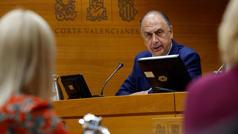 Hombre con traje azul interviene ante una mesa con micrófono en una sala con paneles de madera y el texto Corts Valencianes de fondo.