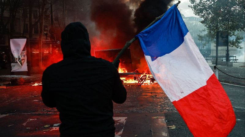 Manifestante de espaldas ondea una bandera francesa frente a una barricada en llamas durante unos disturbios callejeros en una calle adoquinada.