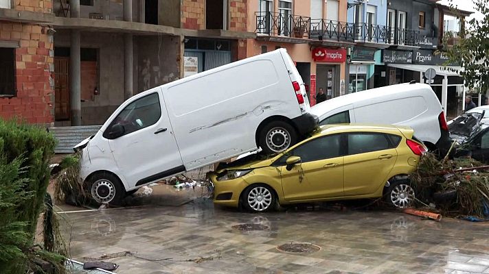 Calle urbana inundada con una furgoneta blanca apilada sobre un coche amarillo, rodeados de lodo, ramas y edificios afectados por una riada.