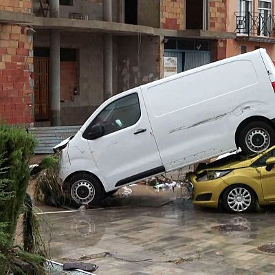 Calle urbana inundada con una furgoneta blanca apilada sobre un coche amarillo, rodeados de lodo, ramas y edificios afectados por una riada.