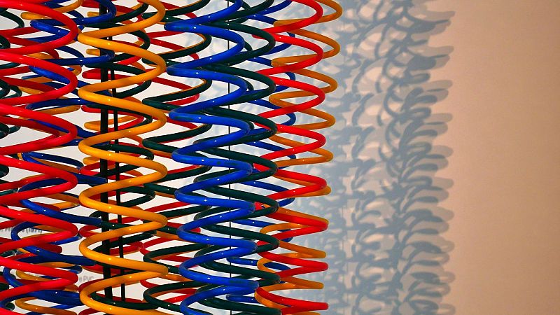 Instalaci�n art�stica de resortes verticales de colores vivos que proyectan sombras onduladas sobre una pared clara.