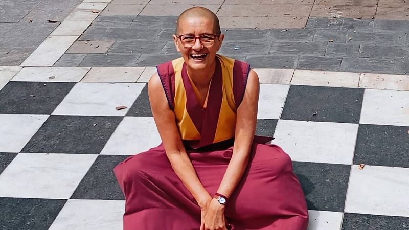 Marina Rizzo, con cabeza rapada, gafas rojas y hábito budista tibetano, sonríe sentada sobre un tablero de ajedrez exterior.