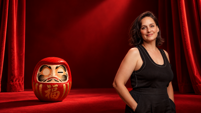 La actriz Patricia Estremera posa sonriente junto a un muñeco daruma japonés en un escenario iluminado con tonos rojos y cortinajes de terciopelo.