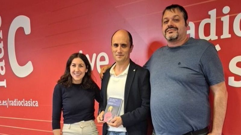 Tres personas posan sonrientes ante un fondo rojo corporativo en un estudio de radio, mostrando un libro y ambiente cordial.
