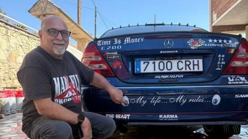 Hombre sonriente agachado junto a un coche sed�n azul oscuro decorado con m�ltiples pegatinas y frases personalizadas en el maletero.