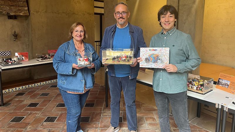 Tres personas posan sonrientes en una sala de exposiciones mostrando vitrinas y un catálogo con dioramas en miniatura.