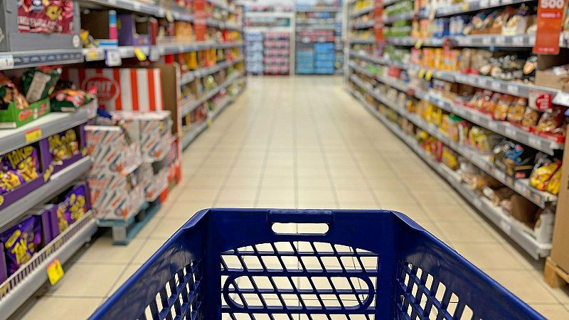 Vista en primera persona desde un carro de compra en el pasillo de un supermercado, flanqueado por estanter�as llenas de productos.