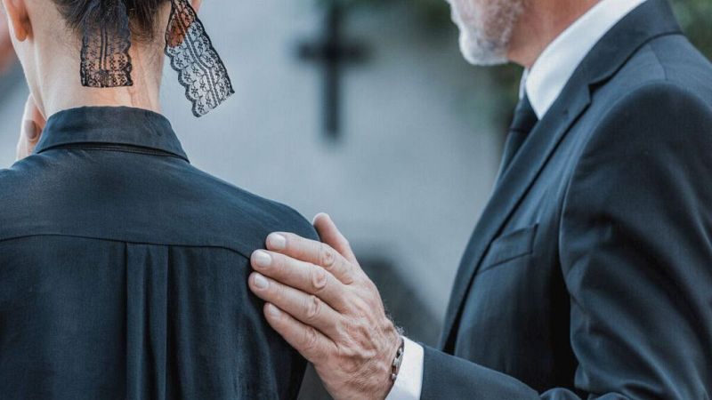 Un hombre mayor con traje oscuro consuela a una mujer vestida de negro con velo de encaje, apoyando su mano sobre el hombro de ella.