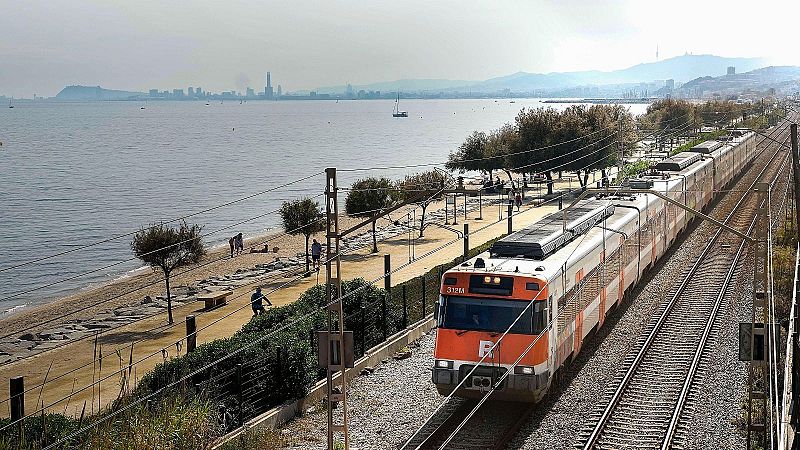 Un tren de cercan�as circula por la v�a junto a la playa del Maresme, con un ciclista en el paseo mar�timo y el mar de fondo bajo un cielo despejado.