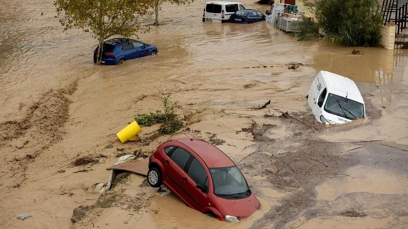 Vista en picado de una zona urbana inundada con agua y fango, donde varios coches y furgonetas aparecen atrapados y parcialmente sumergidos.