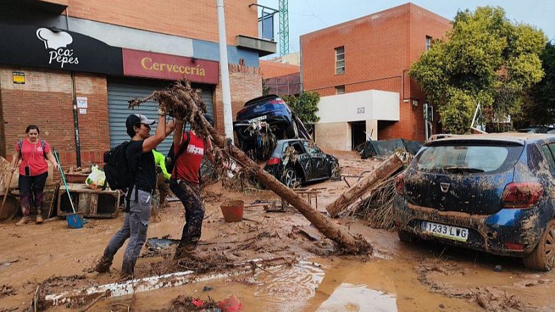 Dos voluntarios retiran un tronco cubierto de lodo en una calle afectada por inundaciones en Valencia, junto a vehículos sepultados y edificios con negocios locales.